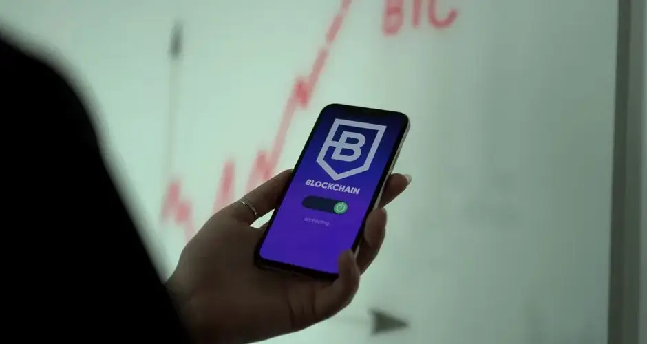 A close-up of a hand holding a smartphone displaying a blockchain app with a stock graph in the background.