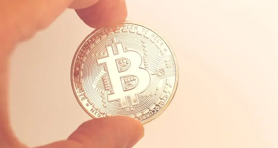 A close-up image of a hand holding a Bitcoin coin against a neutral background.