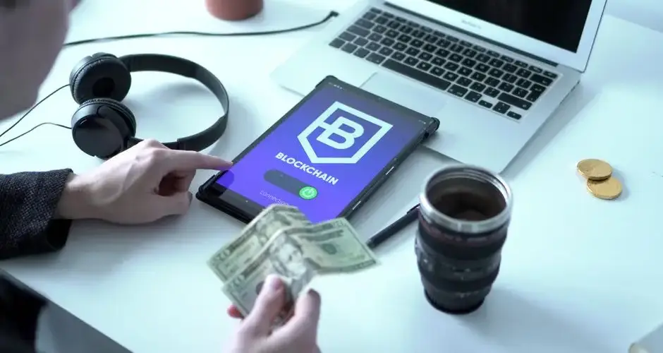 Tablet with blockchain app, laptop, and dollar bills on desk depicting digital finance.