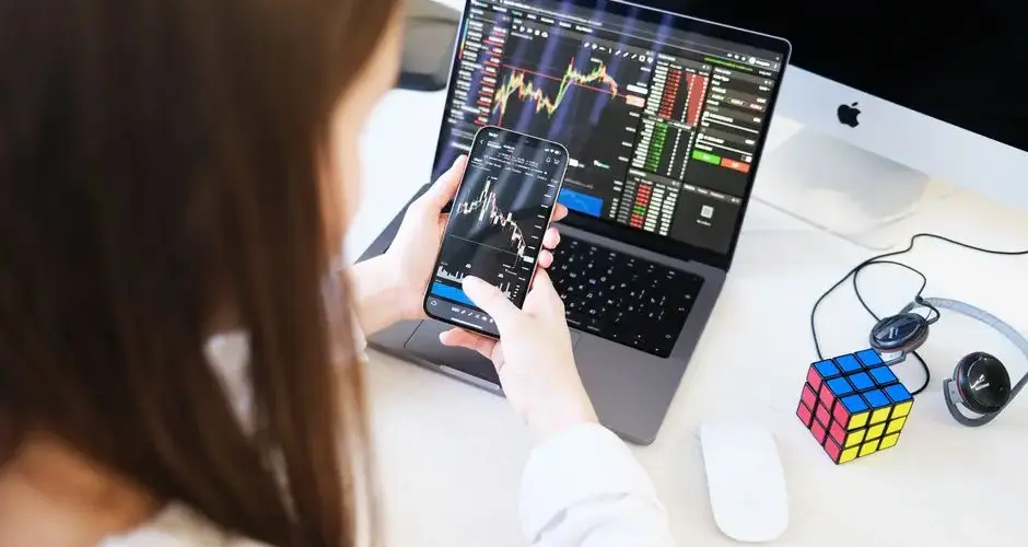 A woman trading cryptocurrency with charts on a smartphone and laptop, showcasing digital finance technology.