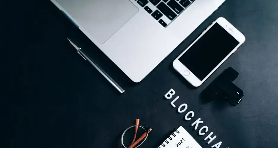 Overhead shot of a desk with a laptop, smartphone, calendar, pen, and 'BLOCKCHAIN' text.