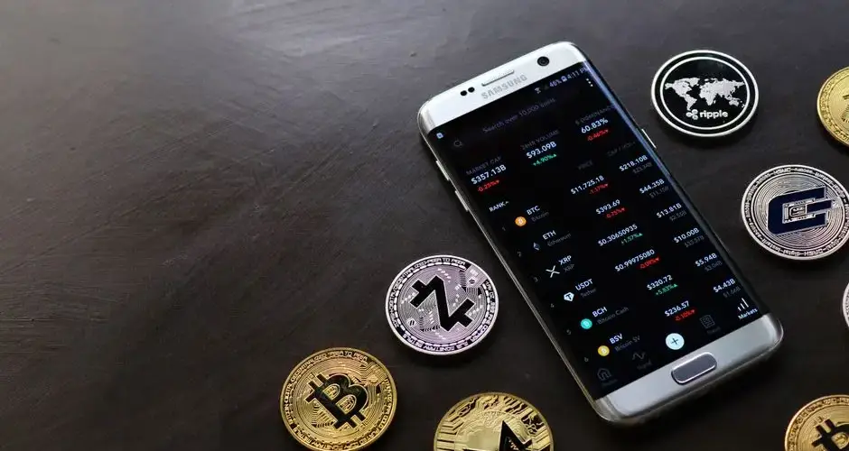A collection of cryptocurrency coins and a smartphone displaying a digital trading platform.