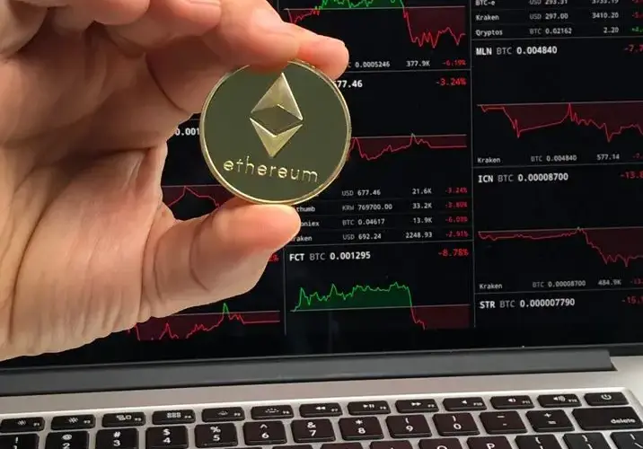 A hand holds an Ethereum coin in front of a laptop displaying cryptocurrency trading charts.