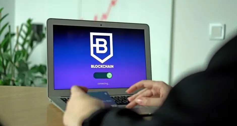 Hand holding a card near a laptop displaying blockchain connection screen.