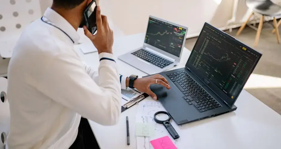 Man holding phone, working on multiple laptops with financial charts at desk.