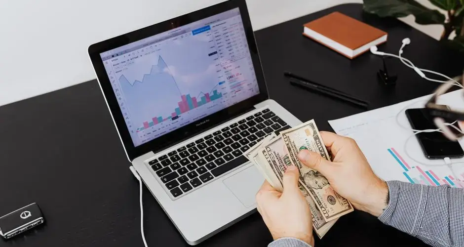 Person counting dollar bills with financial graphs on a laptop screen, symbolizing financial analysis and planning.