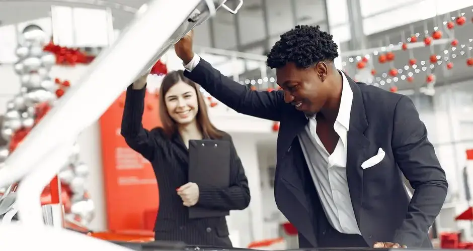 Positive smiling diverse stylish man and female dealer checking under car motor hood and smiling while choosing new car in car showroom