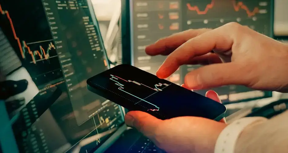 Hands interact with smartphone for analyzing cryptocurrency trading on digital screens.