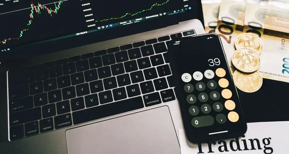Laptop screen with trading charts, calculator app, Euros and Bitcoin coins on desk.