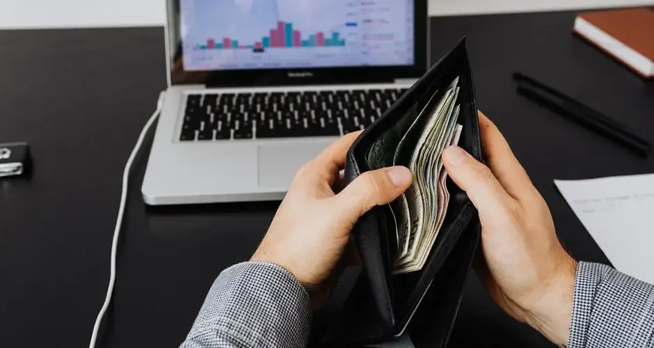 Man holding a leather wallet with bills in front of a laptop displaying financial graphs.