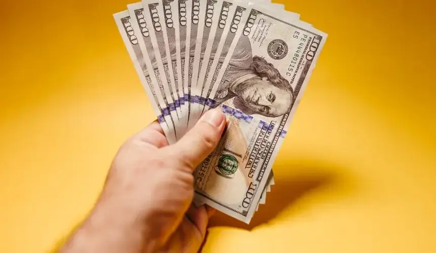 Close-up of a hand holding multiple US dollar banknotes against a yellow background.