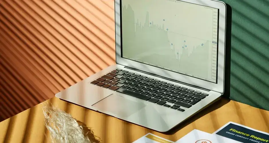 A laptop displaying analytics with financial papers on a textured surface, captured in warm lighting.