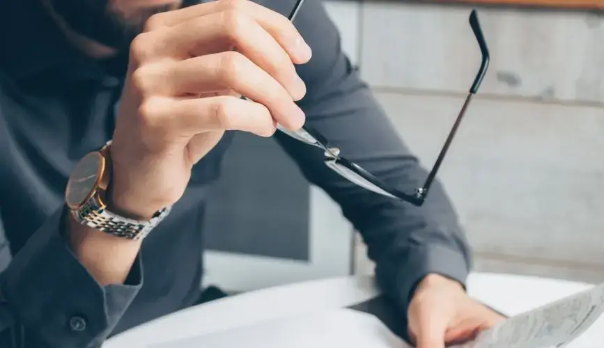 Man in formal attire reviewing paperwork, holding glasses. Business setting.