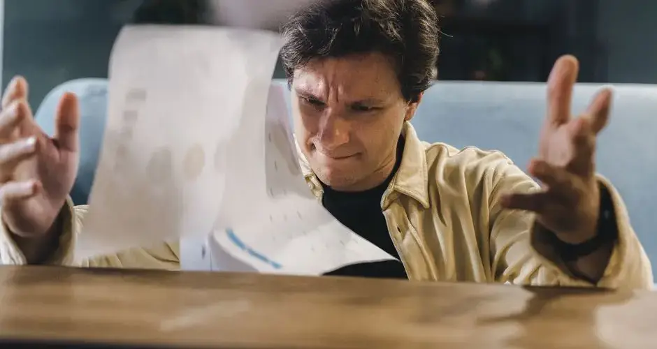 Stressed man throwing documents in frustration at a wooden desk in an indoor setting.