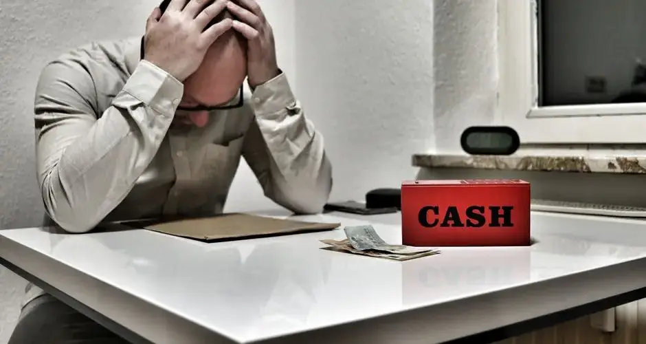 Bald man holding head in despair at desk with cashbox and money. Indoor office stress concept.