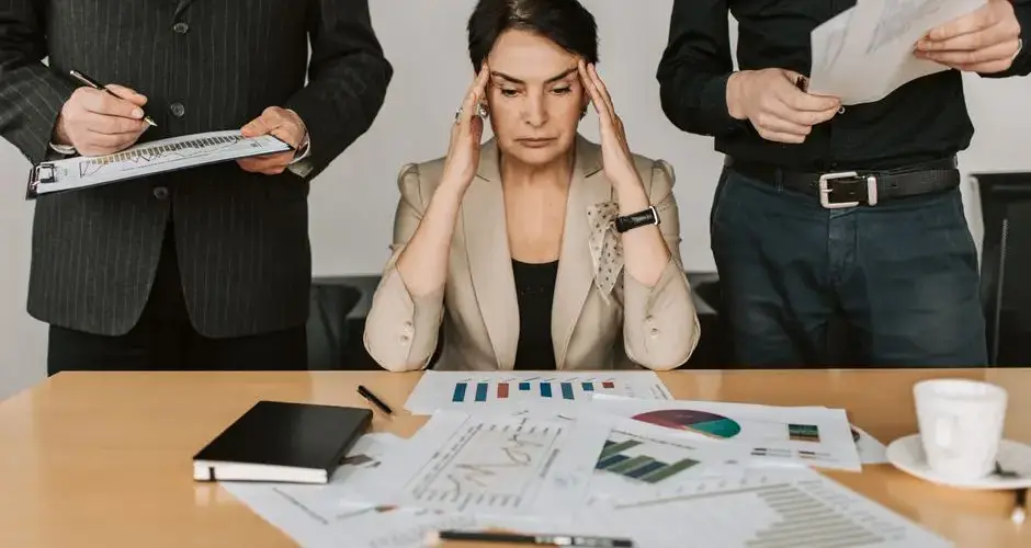A stressed businesswoman holding her head with colleagues holding documents in a corporate office setting.