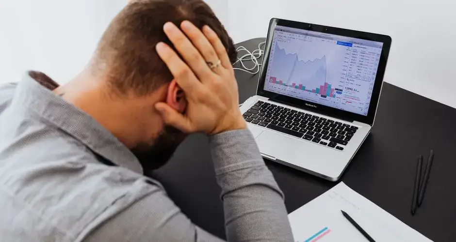 Stressed man at desk looking at declining stock charts on laptop, indicating financial loss.