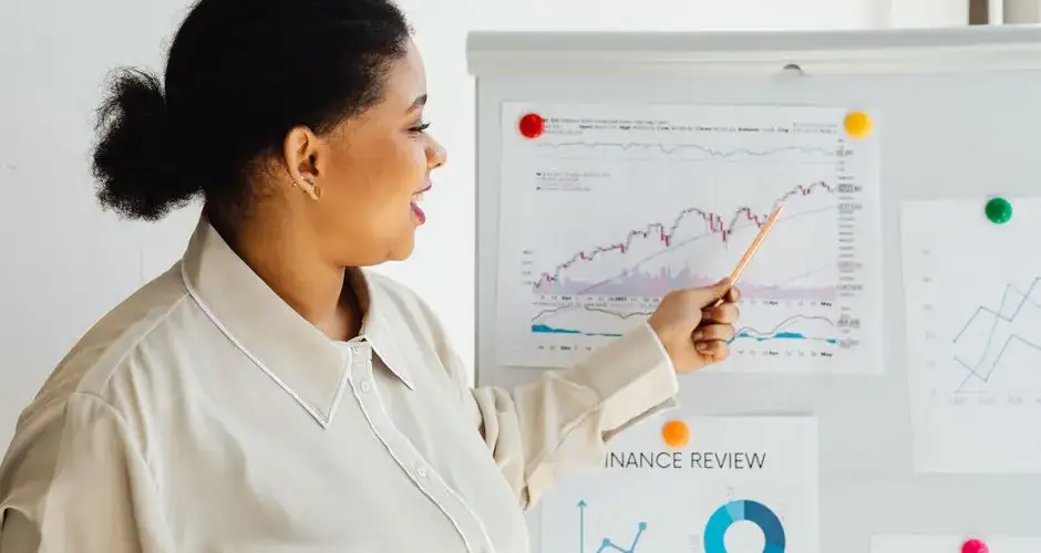A woman points at a stock market chart on a whiteboard, explaining financial trends.