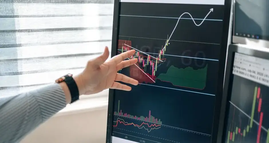 Close-up of a hand pointing at stock market graphs on a monitor in a workspace.
