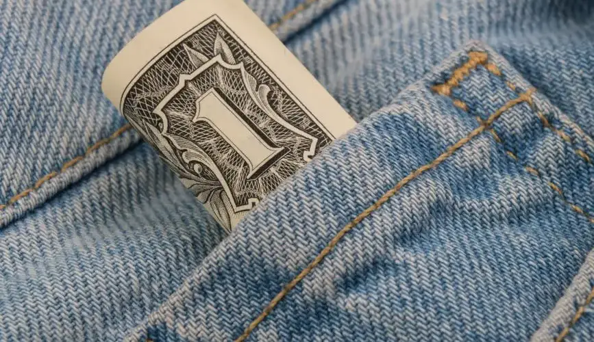 Close-up of a rolled dollar bill tucked into a denim pocket, symbolizing savings and finance.