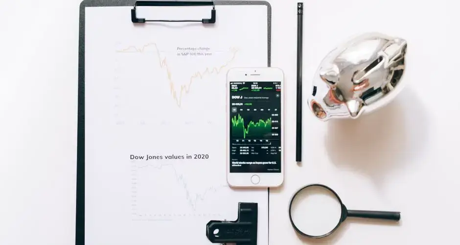 Top view of financial analysis tools including a cellphone, clipboard, magnifying glass, and piggy bank on a white desk.