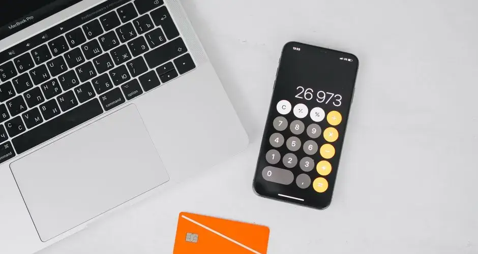 A laptop, smartphone displaying calculator, and credit card on a white table.