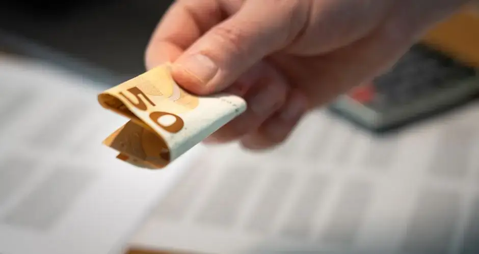 A detailed image of a hand holding a folded 50 euro banknote, with documents in the background.