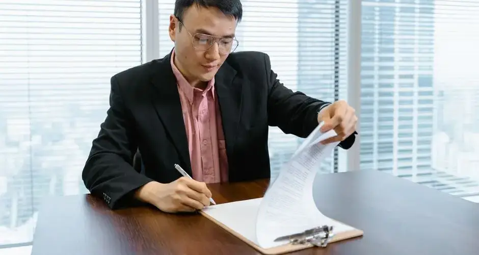 Asian businessman in formal wear signs paperwork at office desk.