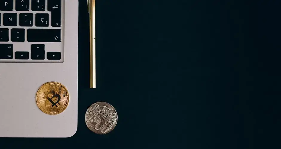 Close-up view of Bitcoin and Ethereum coins next to a laptop and pen on a black surface.