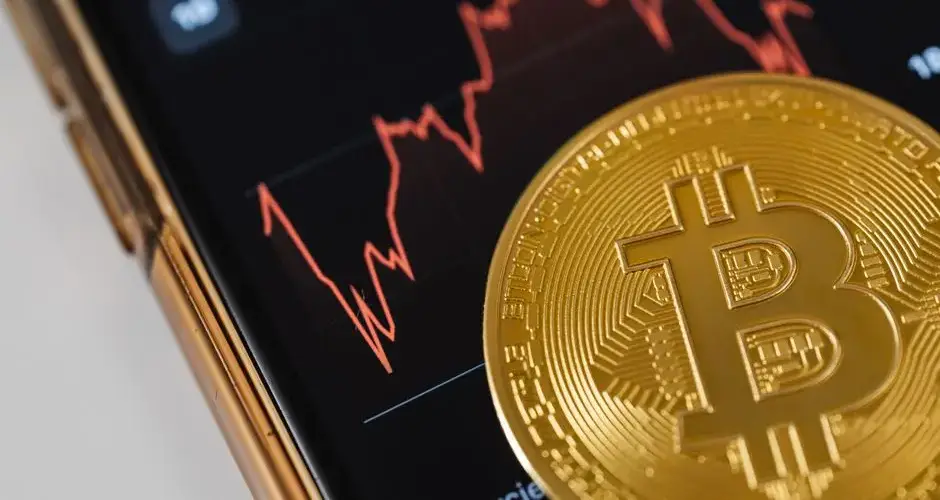 A gold Bitcoin coin on a smartphone displaying a fluctuating stock market chart.