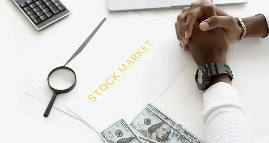 Hands folded over stock market documents with calculator and cash on desk.