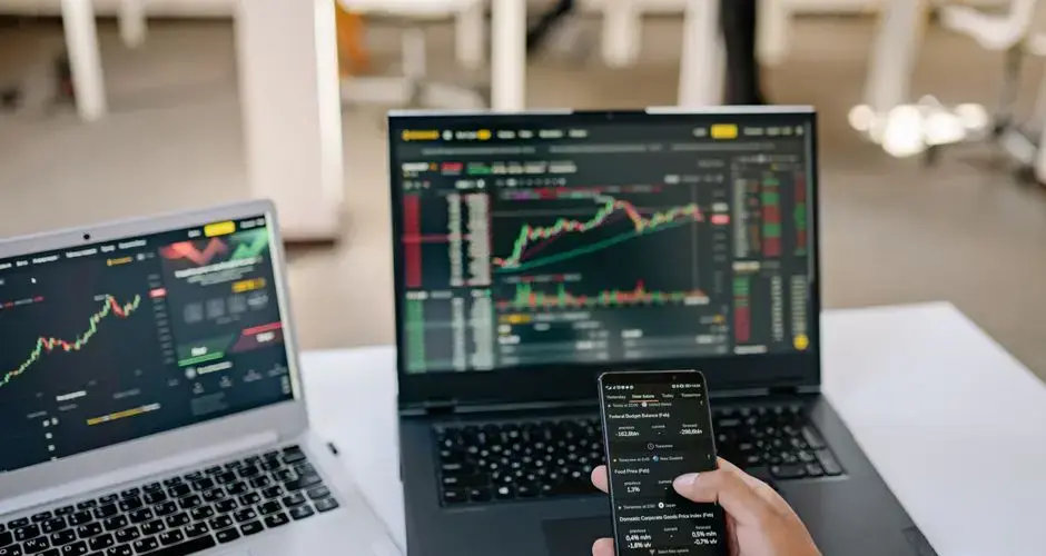 A professional tracks stock market trends using two laptops and a smartphone, showcasing digital finance tools.