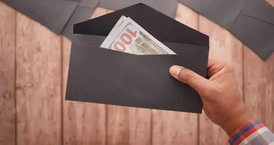 A hand holds a black envelope containing a 100 dollar bill on a wooden surface.