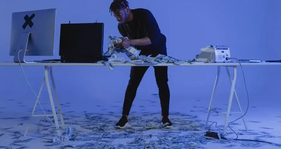 A man counting cash surrounded by computers and money in a blue-lit room, symbolizing cybercrime.