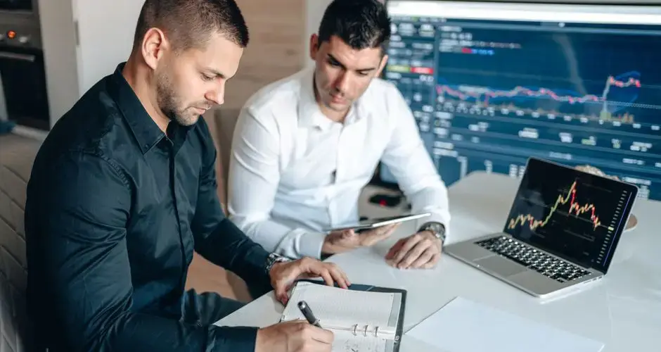 Two businessmen analyzing financial data with digital devices and charts in an office setting.