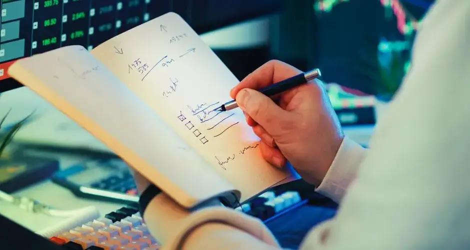 A businessman writes notes in a notebook while analyzing stock market data on a computer screen.