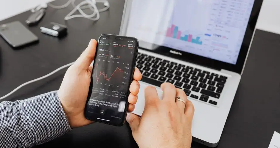Person analyzes stock data with a smartphone and laptop, indoors.