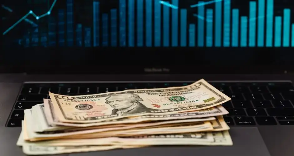 US dollar bills on laptop with financial graph in background, symbolizing wealth and technology.