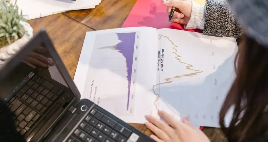 Person reviewing financial graphs and charts while taking notes beside a laptop.