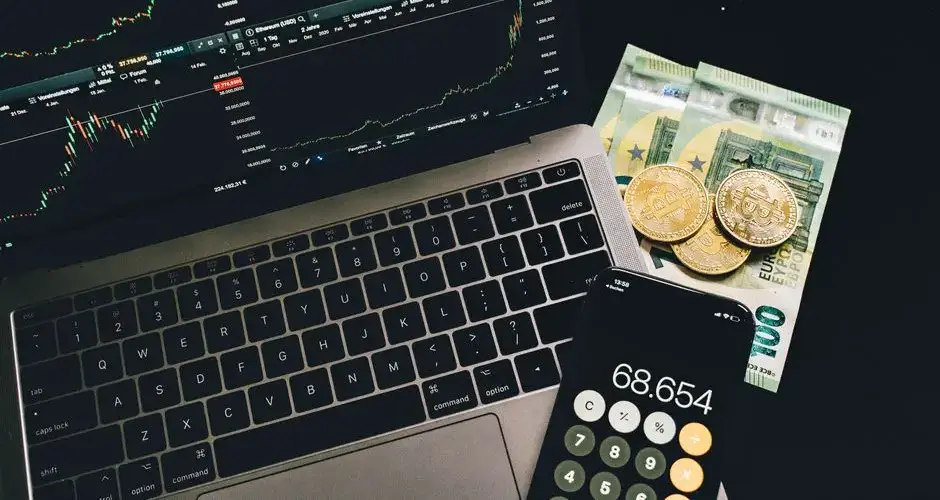 A laptop displaying stock charts with Bitcoin, Euros, and a cellphone calculator, showcasing financial analysis.