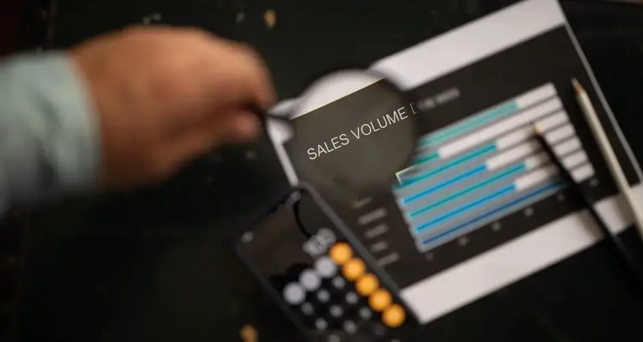 A focused examination of sales volume data using a magnifying glass and calculator.