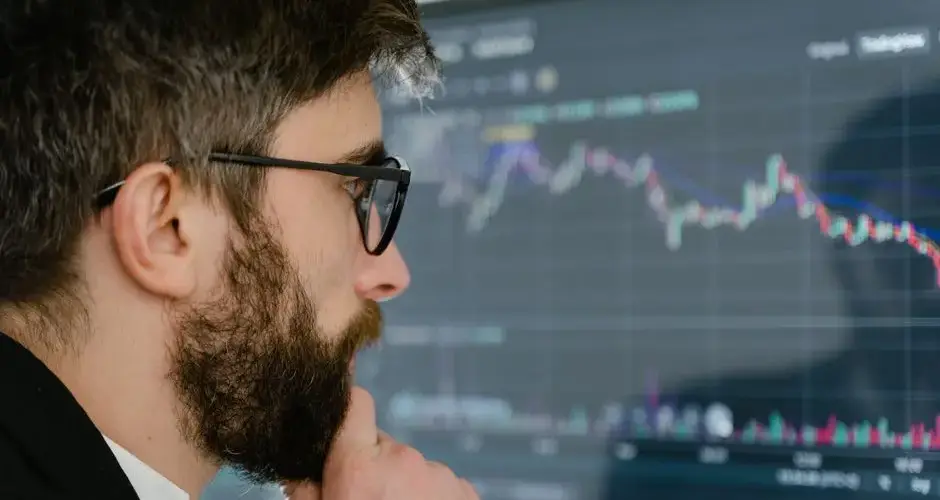 A focused man with glasses studies stock market graphs on a screen, pondering insights.