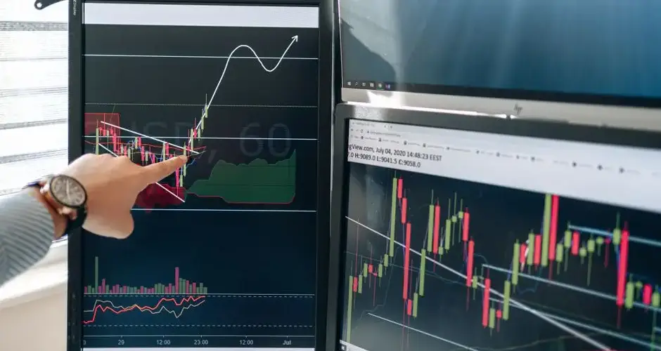 Person pointing at stock market graphs on dual monitors in a modern workspace.
