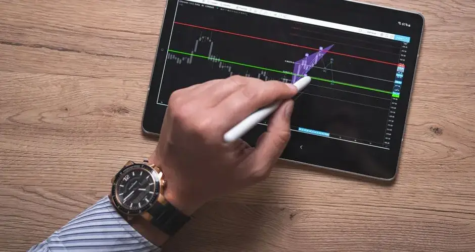 Close-up of a hand using a stylus on a digital trading app on a tablet indoors.