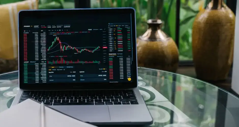Laptop displaying cryptocurrency stocks and graphs on a glass table with a notepad.