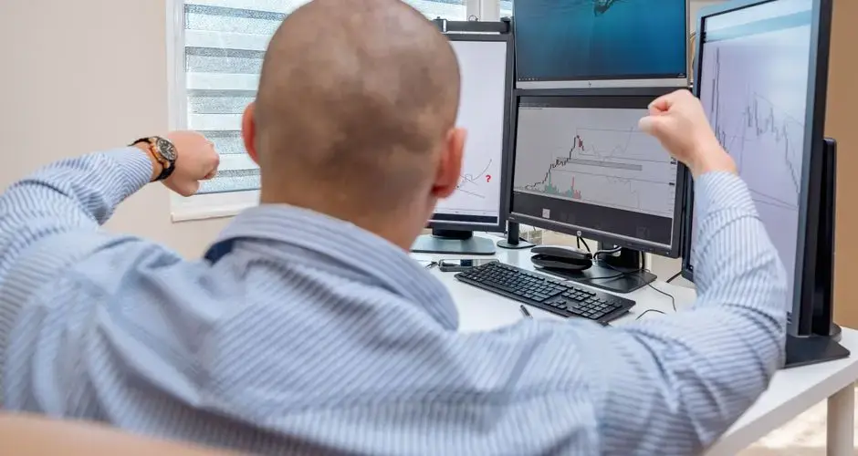 A man celebrates success at a multi-monitor workstation while analyzing stock charts.