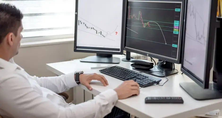A businessman at his desk analyzing financial charts on multiple monitors.