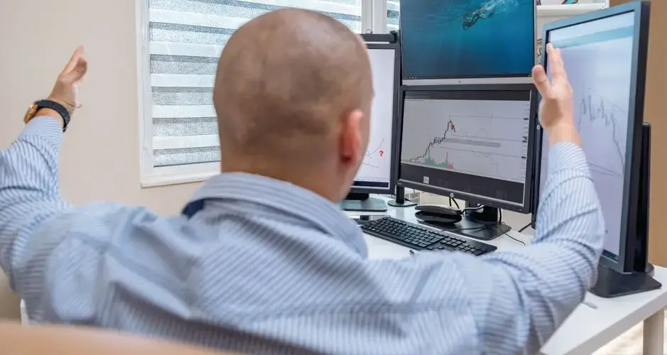 A stock trader in an office raises his hands in celebration while monitoring multiple screens with financial charts.