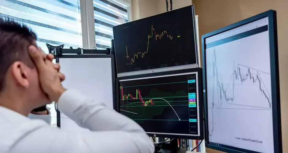 Frustrated man monitoring multiple trading graphs on computer screens in an office setting.