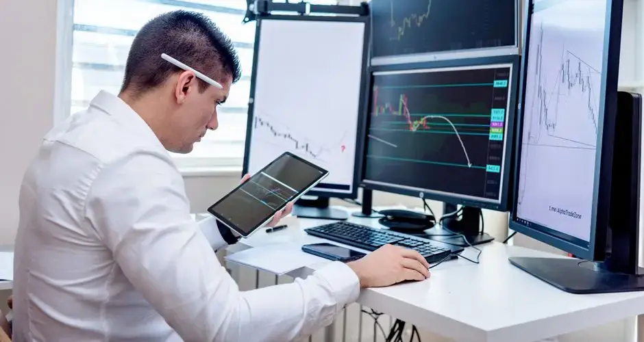 Professional analyzing stock market graphs on multiple monitors at work desk.